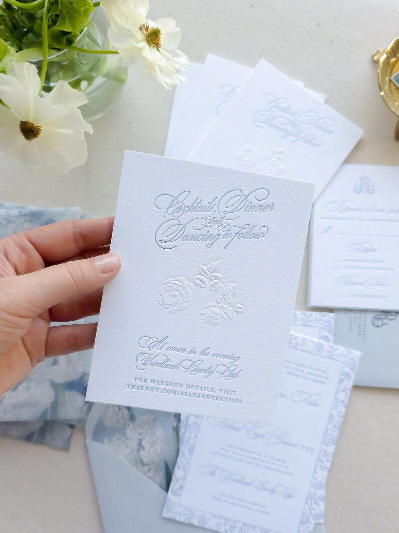 White wedding detail card with sculpted-embossed roses and pale blue script, held above a coordinating invitation suite